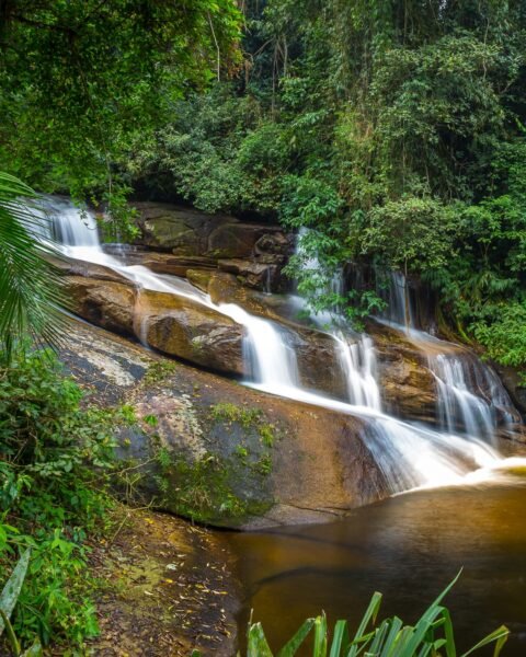 Cachoeira em meio à floresta densa, com água corrente sobre rochas e vegetação tropical ao redor.