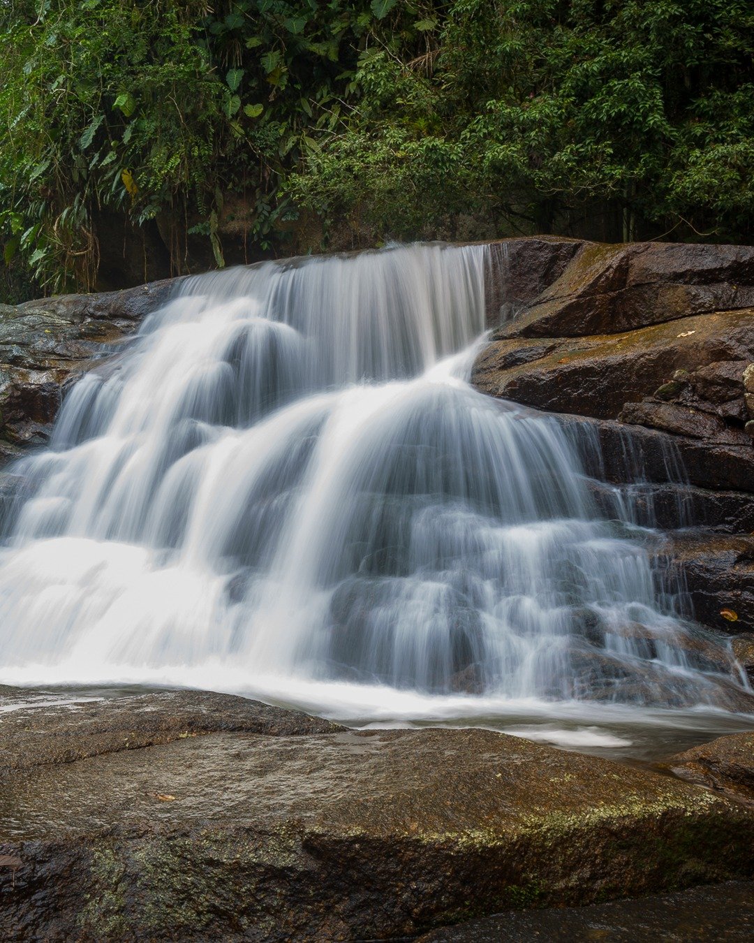 Cachoeira em floresta com fluxo de água longo exposto, cercada por rochas e vegetação verde.