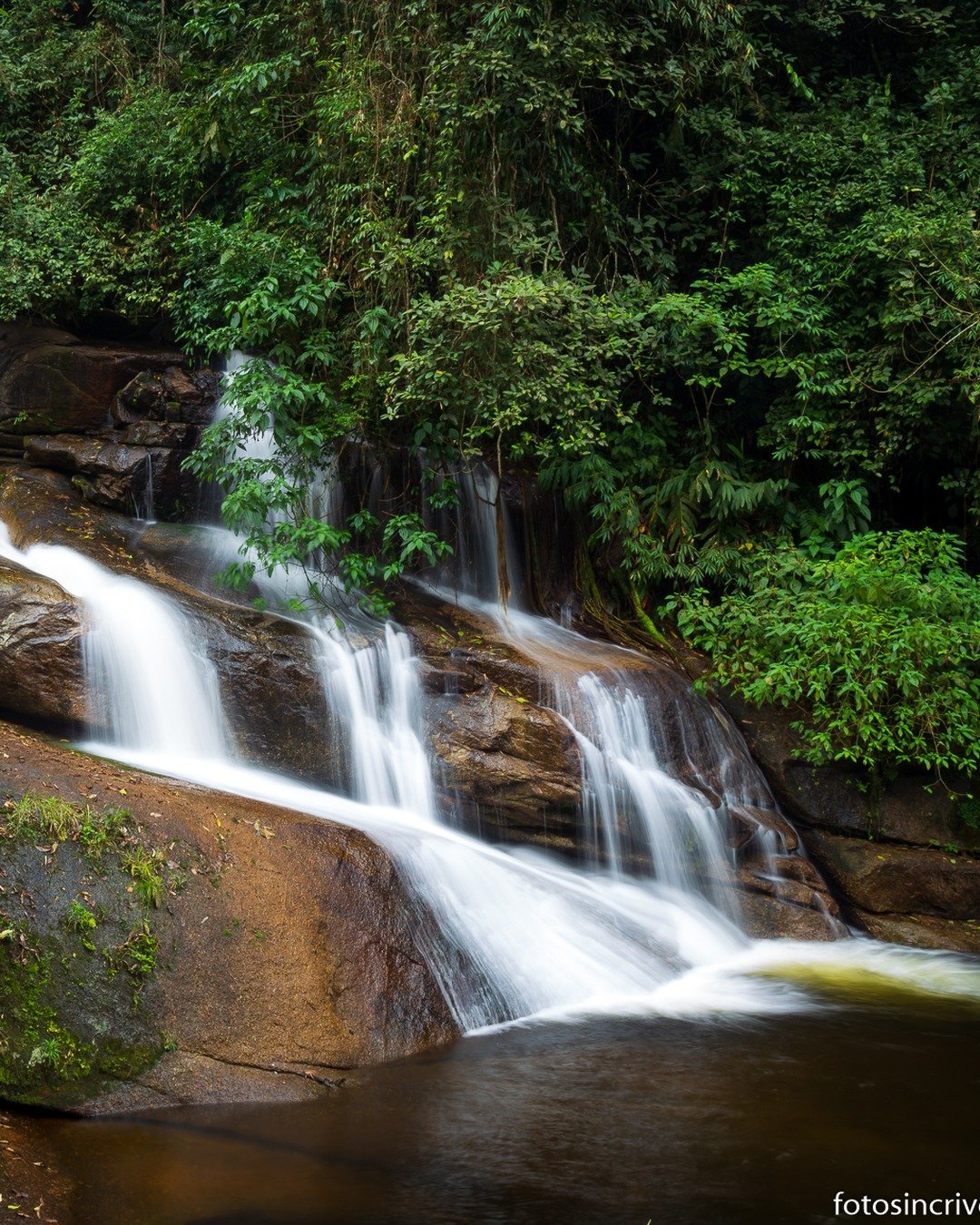 Cachoeira em meio à vegetação tropical, com água descendo por rochas inclinadas e pequena piscina ao fundo.