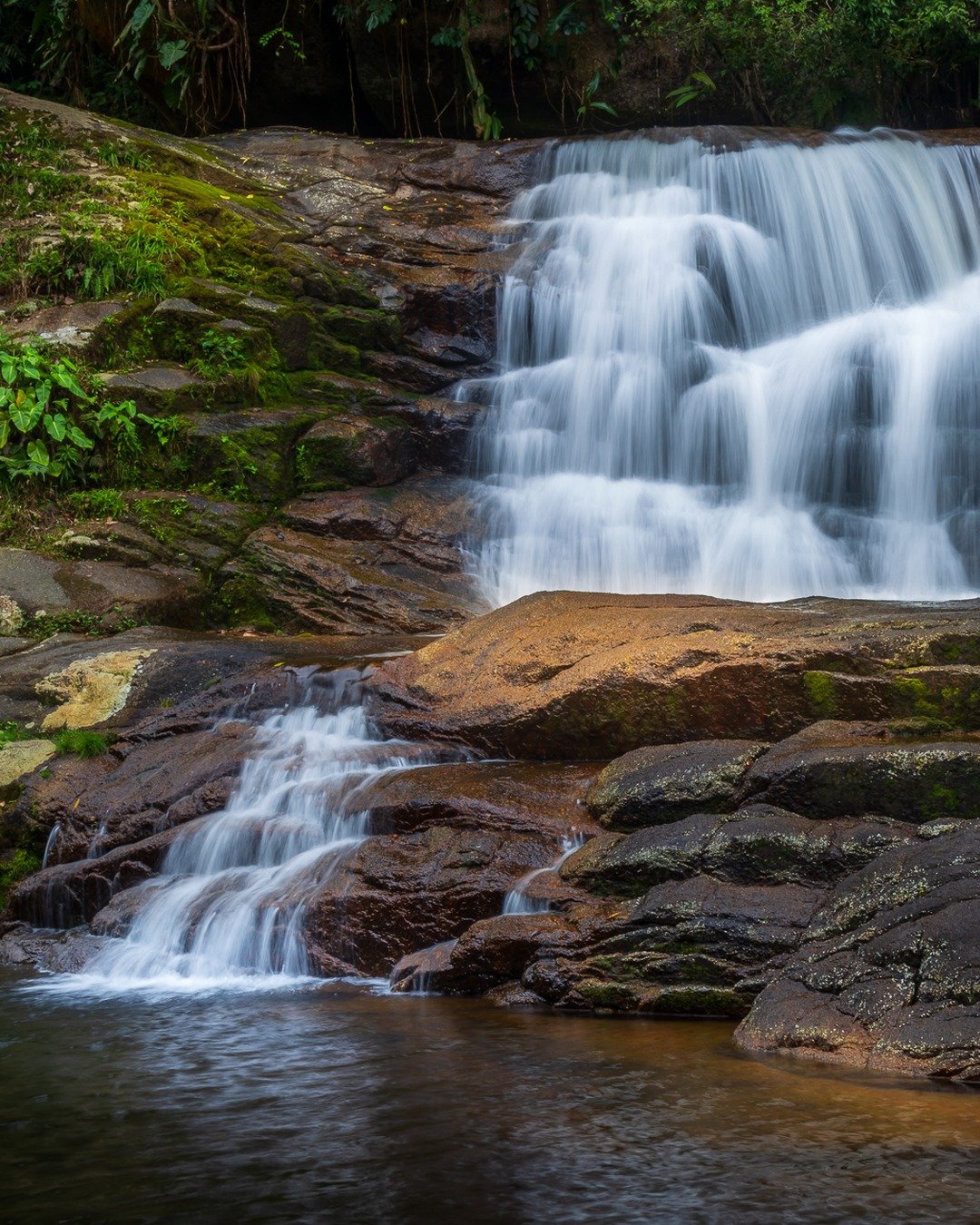 Cachoeira em degraus sobre rochas escuras, cercada por vegetação e com poça de água escura em primeiro plano.