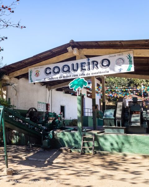 Galpão rústico ao ar livre com faixa 'COQUEIRO' pendurada, máquinas de beneficiamento de café expostas sobre piso de areia e árvores ao redor sob céu azul.