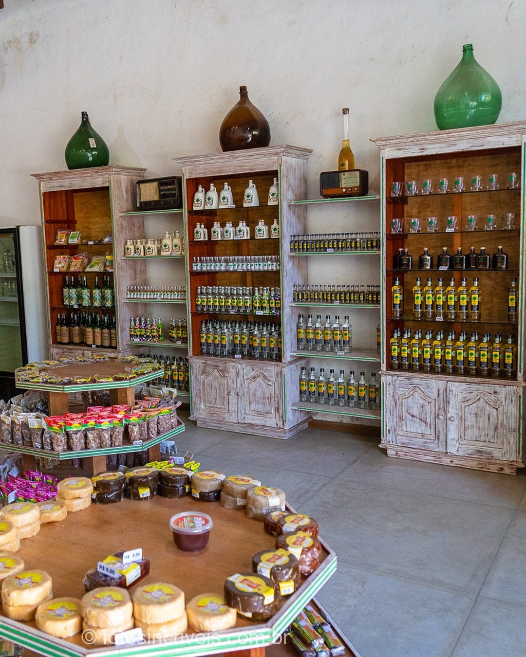 Interior de loja de alambique com prateleiras de madeira rústica repletas de garrafas, licores e potes de doces, além de freezer e balcão de madeira à esquerda.