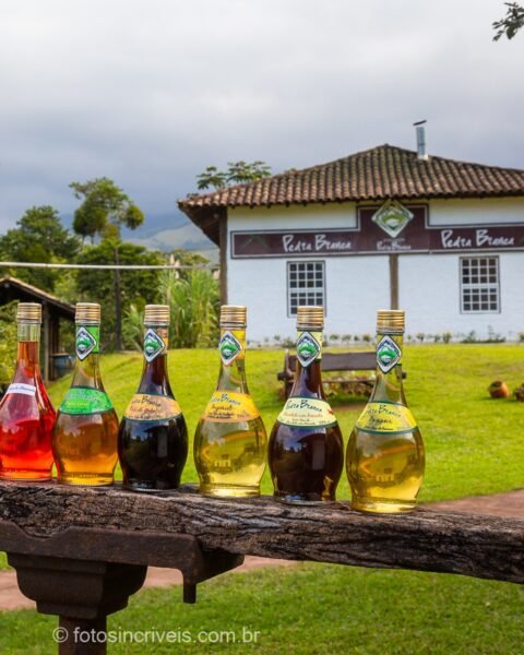 Fileira de garrafas coloridas de licor e cachaça dispostas sobre uma viga de madeira de um alambique, com prédio branco da destilaria 'Péras Brancas' ao fundo, gramado verde, montanhas e céu nublado.