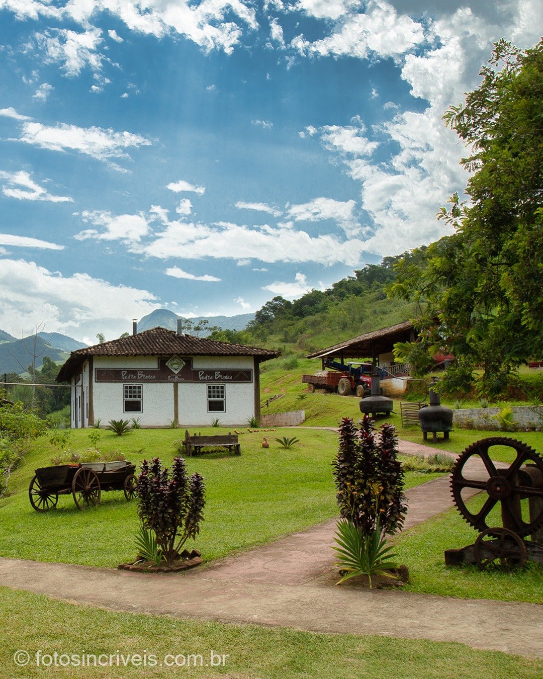 Cena rural com casa colonial branca de telhado de barro, estrutura de madeira com telhado à esquerda, carroça antiga, engrenagem metálica e plantas ornamentais em gramado, sob céu azul com nuvens e montanhas ao fundo.