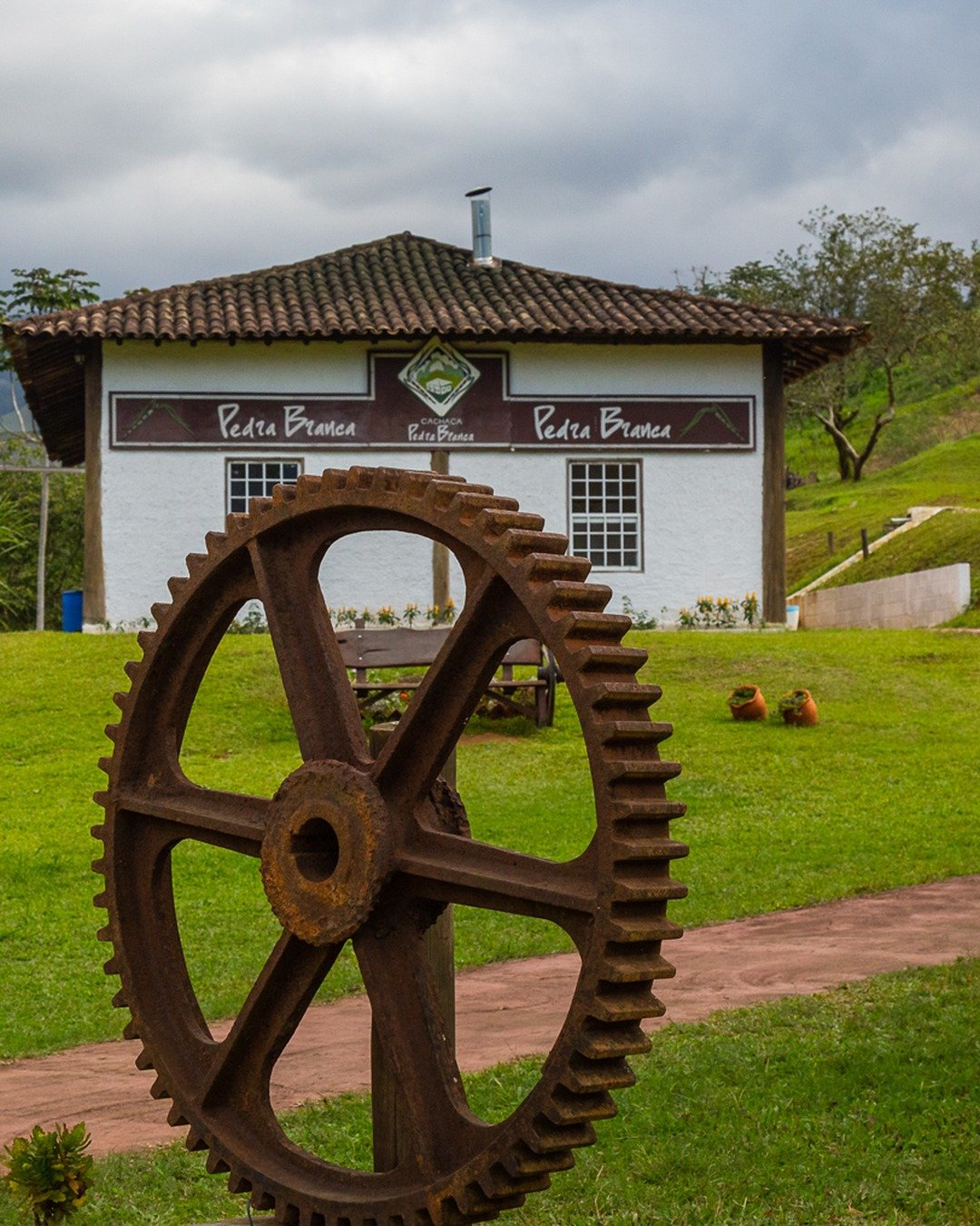 Alambique Pedra Branca: grande engrenagem enferrujada em primeiro plano, casarão branco com placas 'Pedra Branca', gramado verde, caminho de tijolos vermelhos e céu nublado.