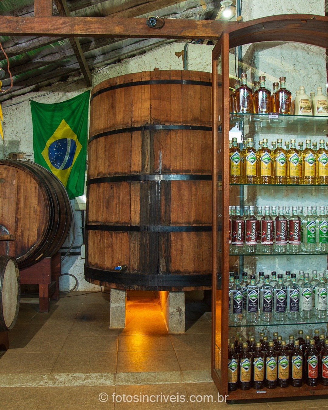 Interior rústico de alambique com barris de madeira, prateleiras repletas de garrafas de licor e bandeira do Brasil ao fundo.
