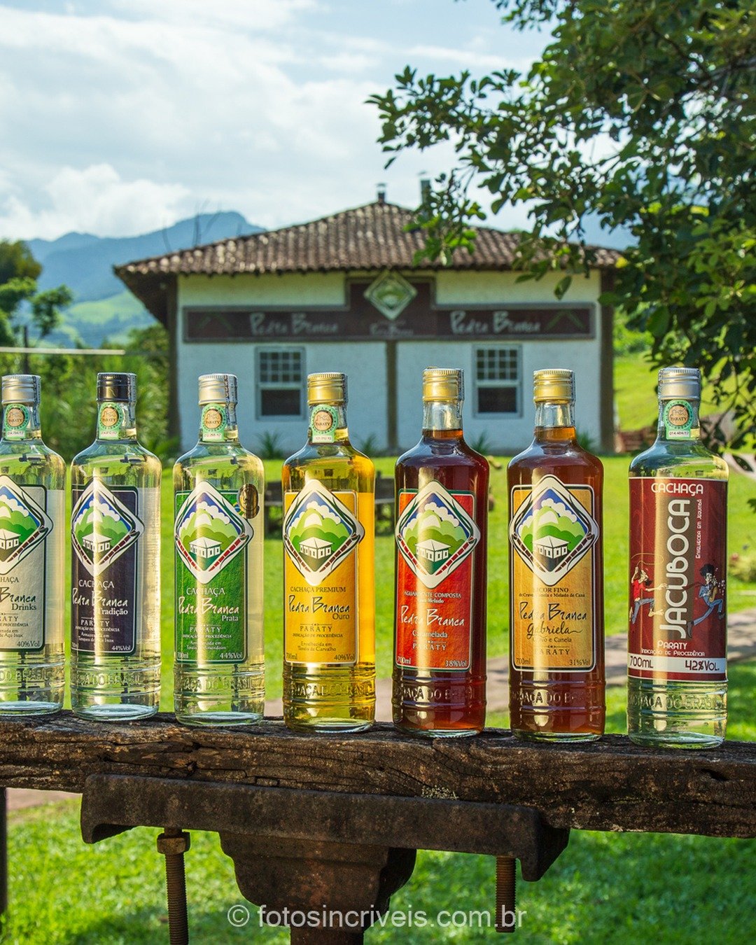 Sete garrafas de cachaça e licores enfileiradas sobre a cerca rústica de madeira, com destilaria de fachada branca e telhado colonial ao fundo, vegetação, carroça antiga e montanhas sob céu azul com nuvens.