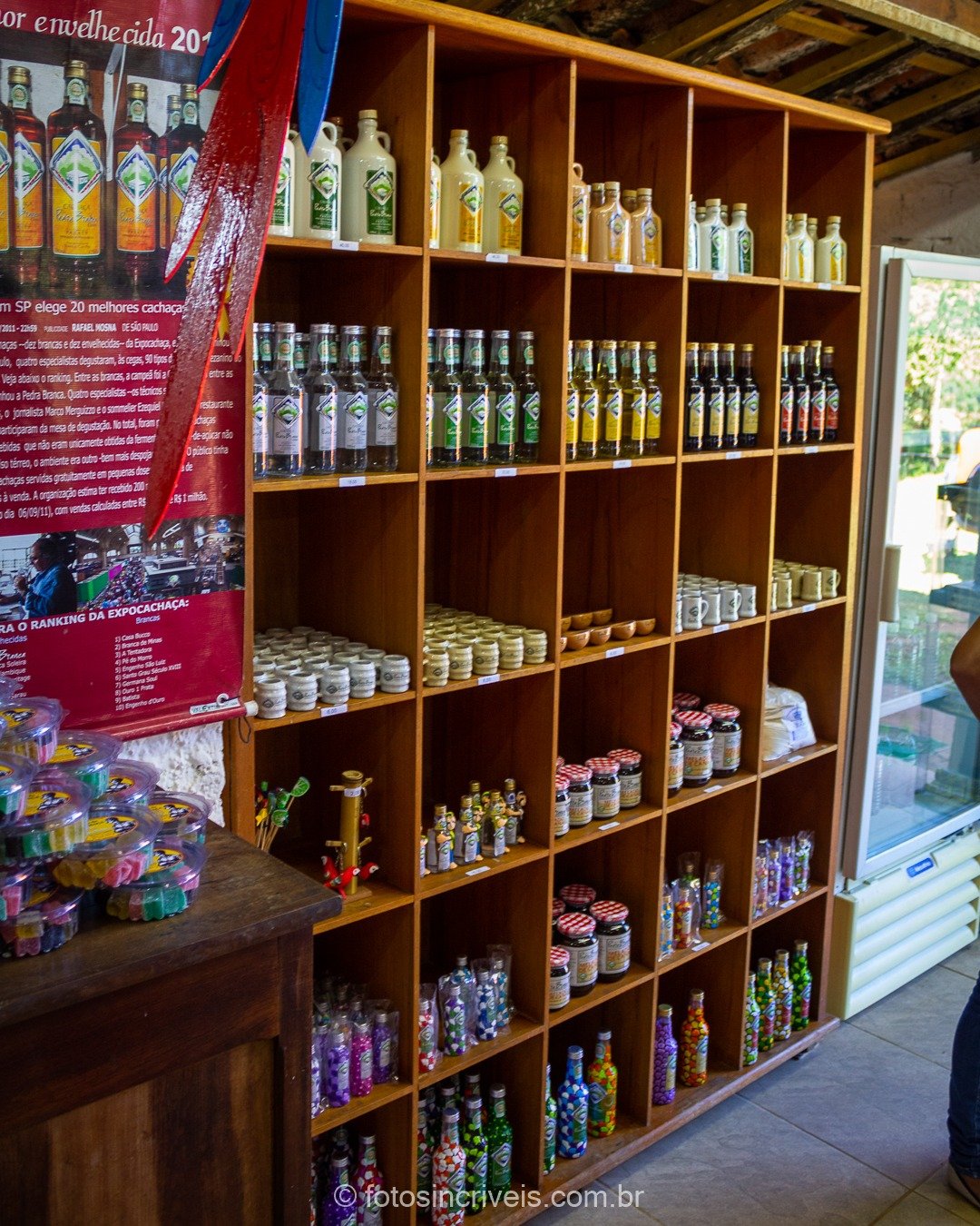 Interior de loja de alambique com prateleiras de madeira repletas de garrafas de cachaça e licores, cartaz vermelho da marca Pedra Branca à esquerda e freezer branco à direita.
