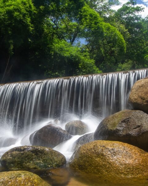 Cachoeira larga com véu d'água sobre rochas, pedras grandes no primeiro plano e mata verde ao fundo sob céu azul com nuvens.
