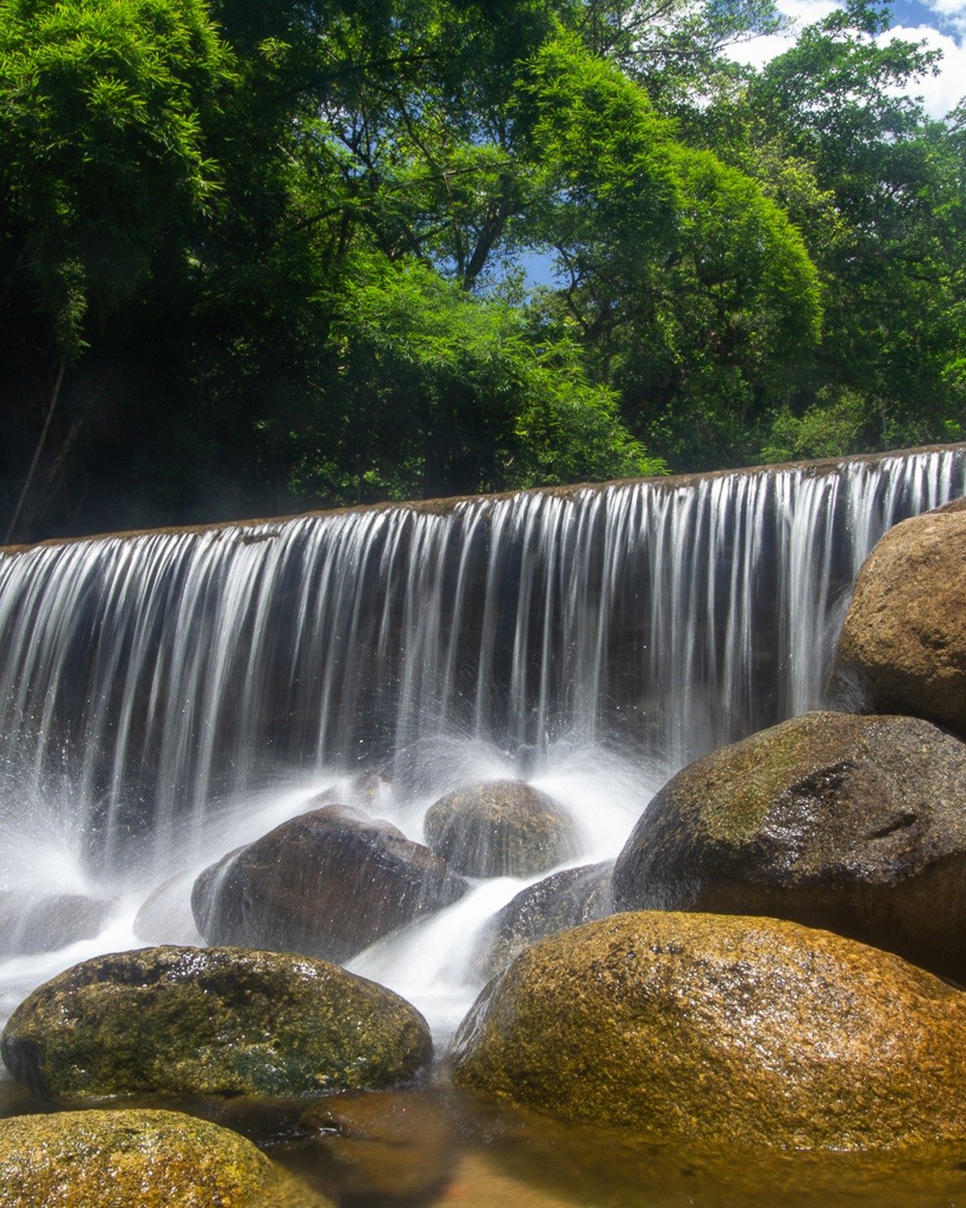 Cachoeira larga com véu d'água sobre rochas, pedras grandes no primeiro plano e mata verde ao fundo sob céu azul com nuvens.