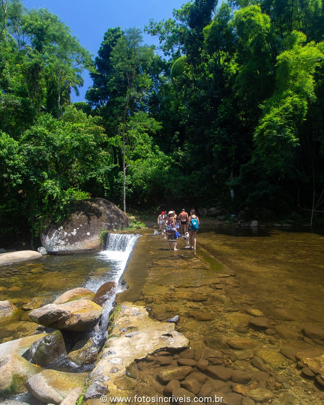 Cachoeira com queda d'água em rio cercado por mata, com várias pessoas banhando-se nas pedras e piscinas naturais.