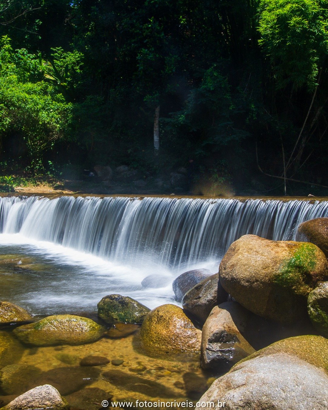Cachoeira larga com cortina de água descendo sobre rochas, cercada por mata atlântica densa e poço raso com pedras no primeiro plano.