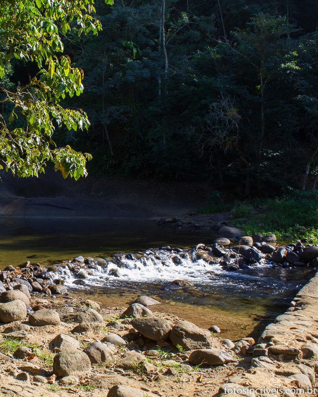 Cena de rio raso e cristalino em meio à mata fechada, com pequena cachoeira sobre rochas à esquerda e um grupo de pessoas caminhando dentro da água no centro da imagem.