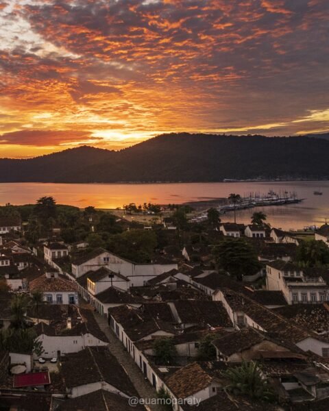 Vista elevada de centro histórico colonial à beira-mar ao pôr do sol, com céu de nuvens alaranjadas sobre morros e água.