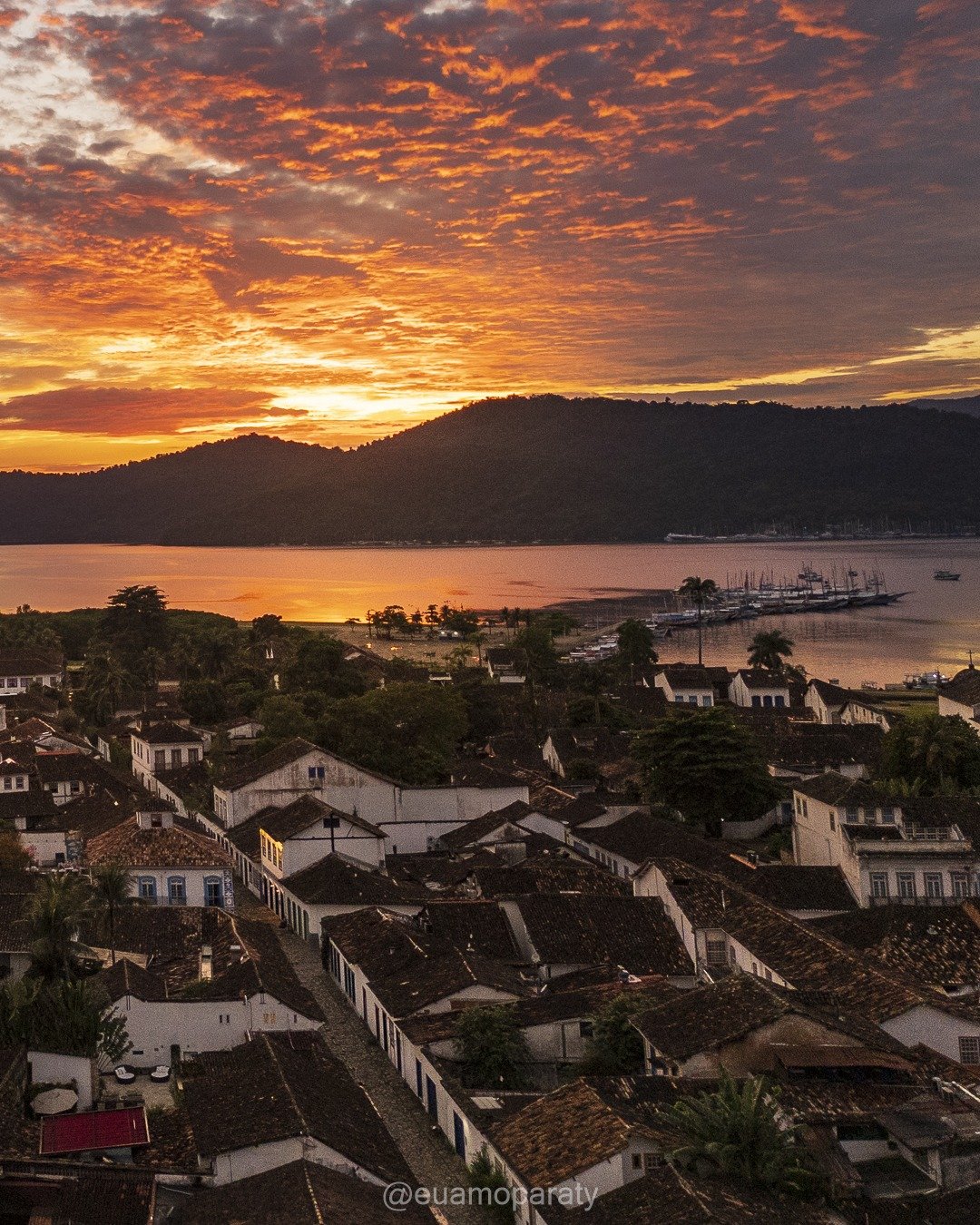 Vista elevada de centro histórico colonial à beira-mar ao pôr do sol, com céu de nuvens alaranjadas sobre morros e água.