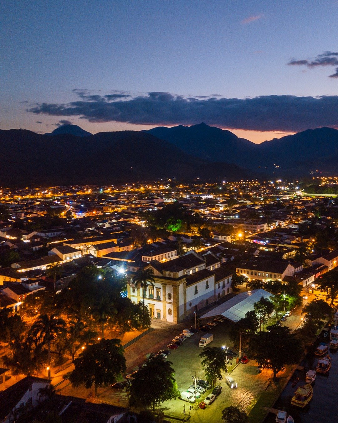 Vista aérea noturna de uma cidade histórica à beira de um rio, com ruas iluminadas, casario colonial, embarcações atracadas e montanhas ao fundo sob céu crepuscular.