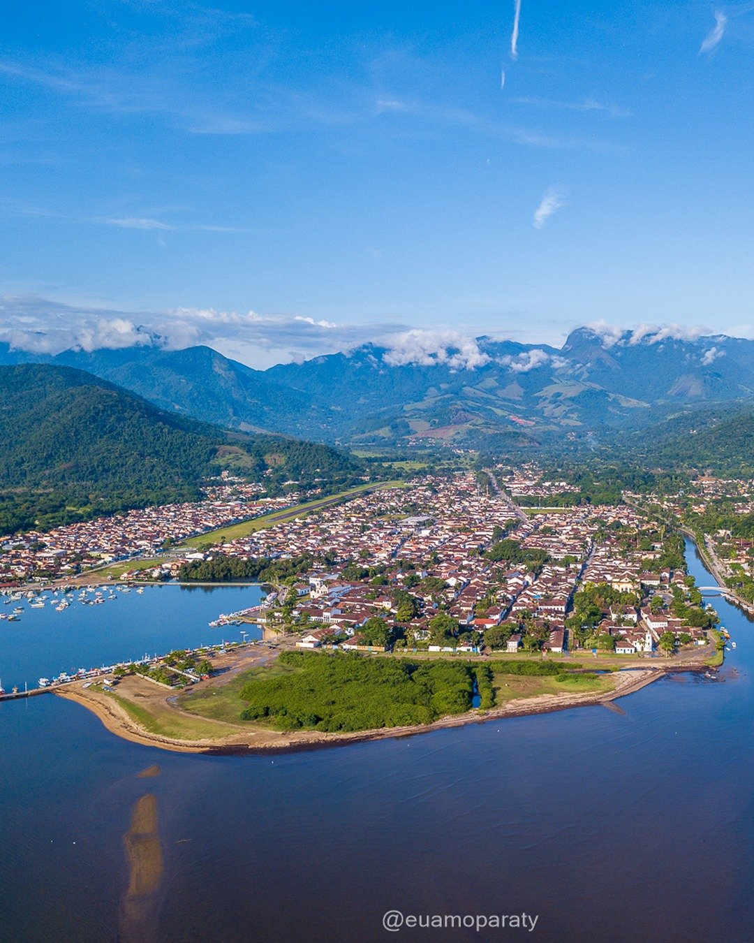 Vista aérea de Paraty com o centro histórico, canal do rio, barcos ancorados, montanhas ao fundo e céu azul com nuvens.