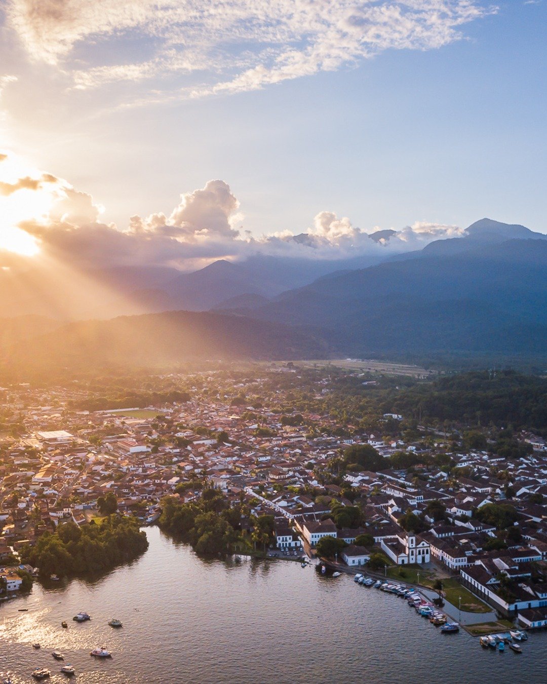 Vista aérea de uma cidade litorânea histórica ao pôr do sol, com raios de sol atravessando nuvens sobre montanhas ao fundo, casario colonial, pista de pouso à esquerda e água com barcos ancorados em primeiro plano.