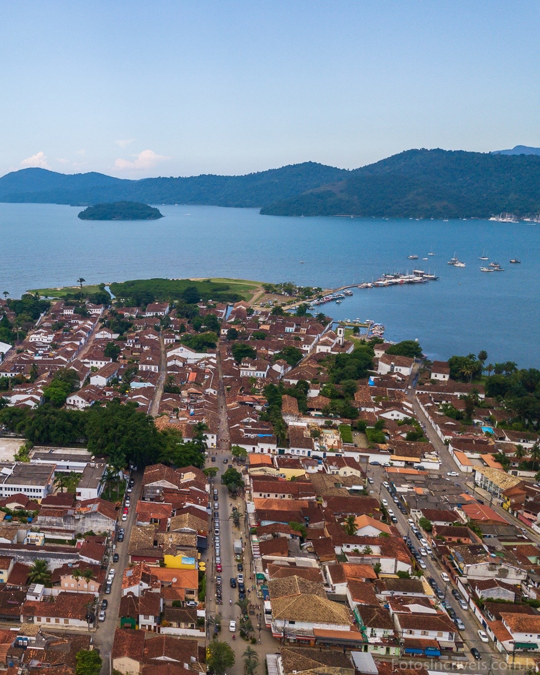 Vista aérea de cidade histórica litorânea com telhados coloniais, rio canal, baía com ilhas e montanhas ao fundo.
