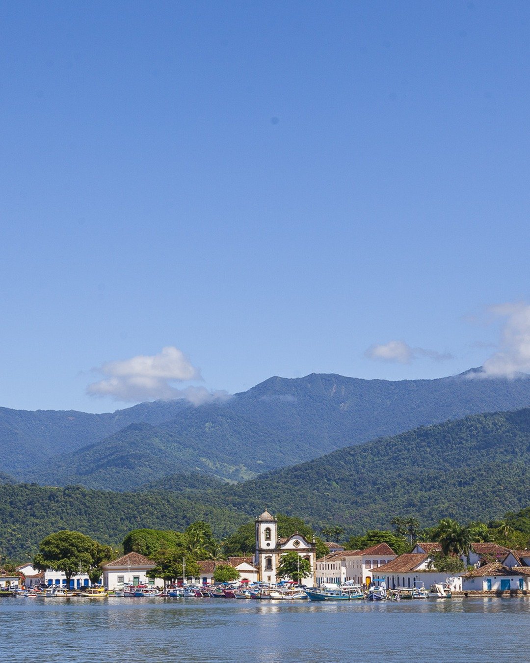 Vista panorâmica de cidade histórica à beira-mar, com igreja, casario colonial, montanhas ao fundo e céu azul.