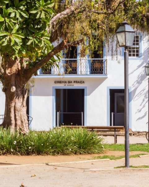 Fachada branca com detalhes azuis do prédio 'Cinema da Praça de Paraty', com varanda no pavimento superior, emoldurada por uma grande árvore à esquerda e um poste de iluminação à direita, sobre gramado e caminho de terra.