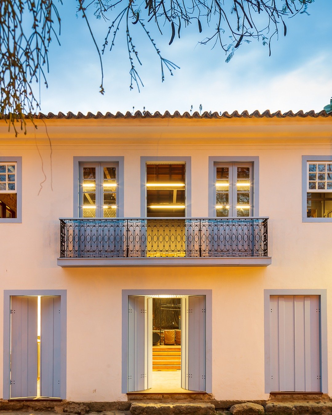Fachada colonial de cor clara com portas e janelas de moldura azul, varanda com gradil de ferro na altura superior e luzes internas acesas ao anoitecer, vista de uma rua de pedra com vegetação ao redor.