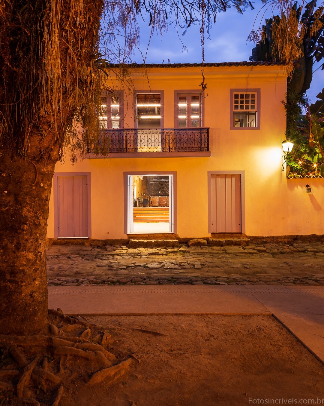 Fachada de casa colonial iluminada por luzes quentes ao anoitecer, com árvore de tronco grosso em primeiro plano à esquerda e rua de pedras à frente, sob céu azul crepuscular.