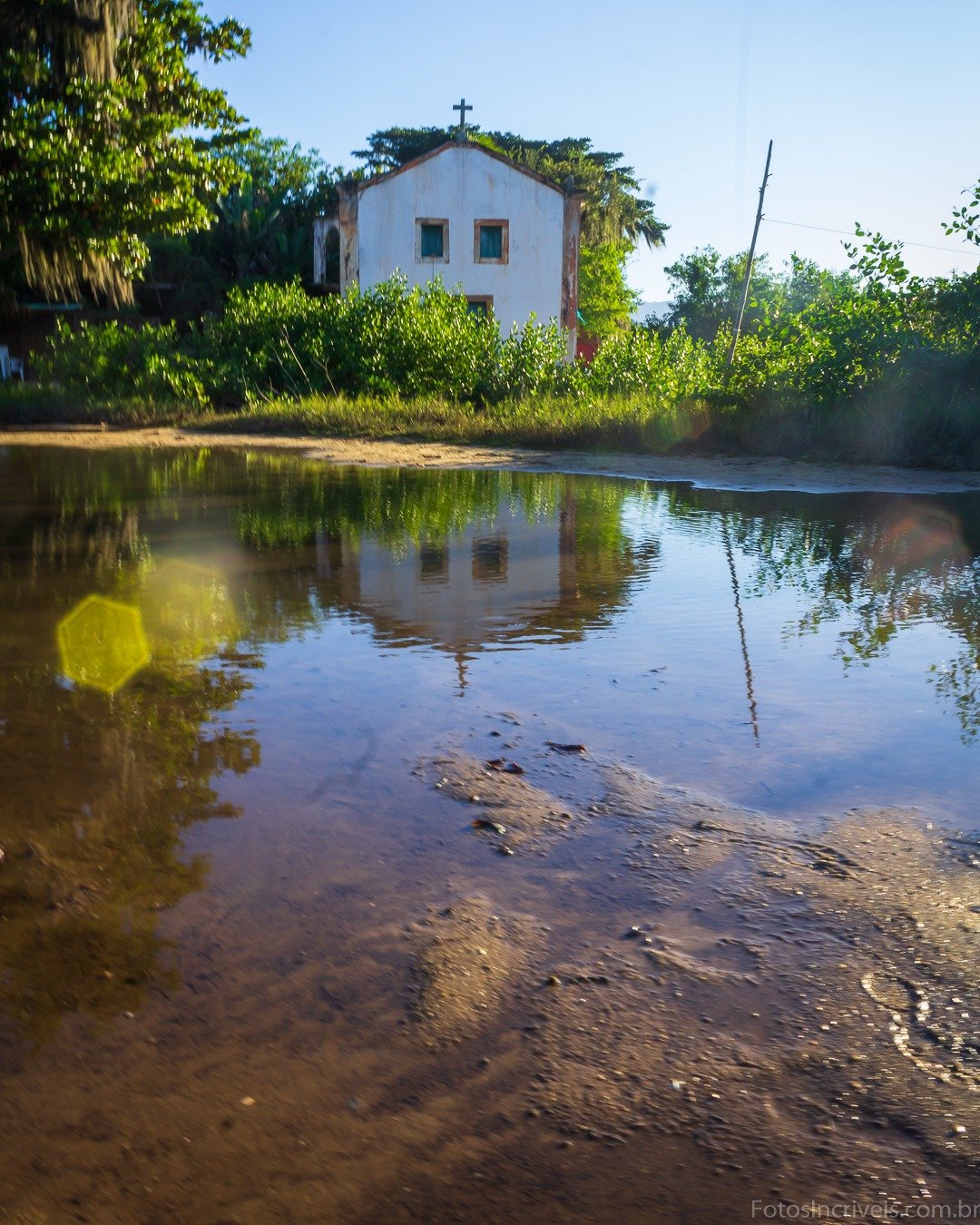 Pequena capela branca com cruz, cercada por vegetação, refletida em uma poça d'água lodosa sob céu azul.