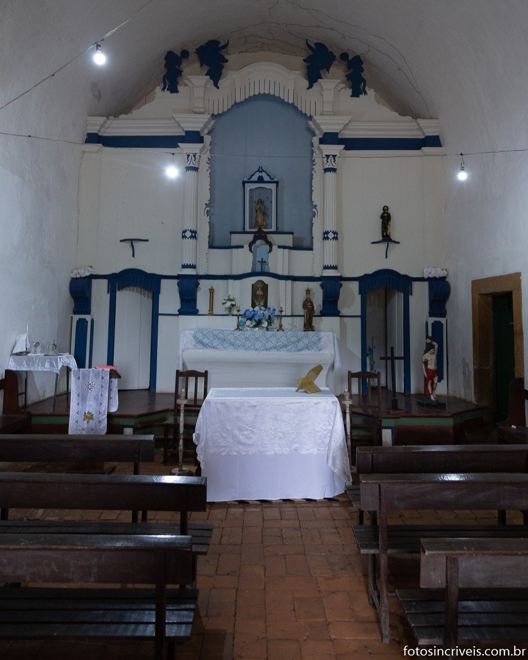Interior de uma pequena igreja, com altar branco e azul ao fundo, bancos de madeira e piso de tijolos.
