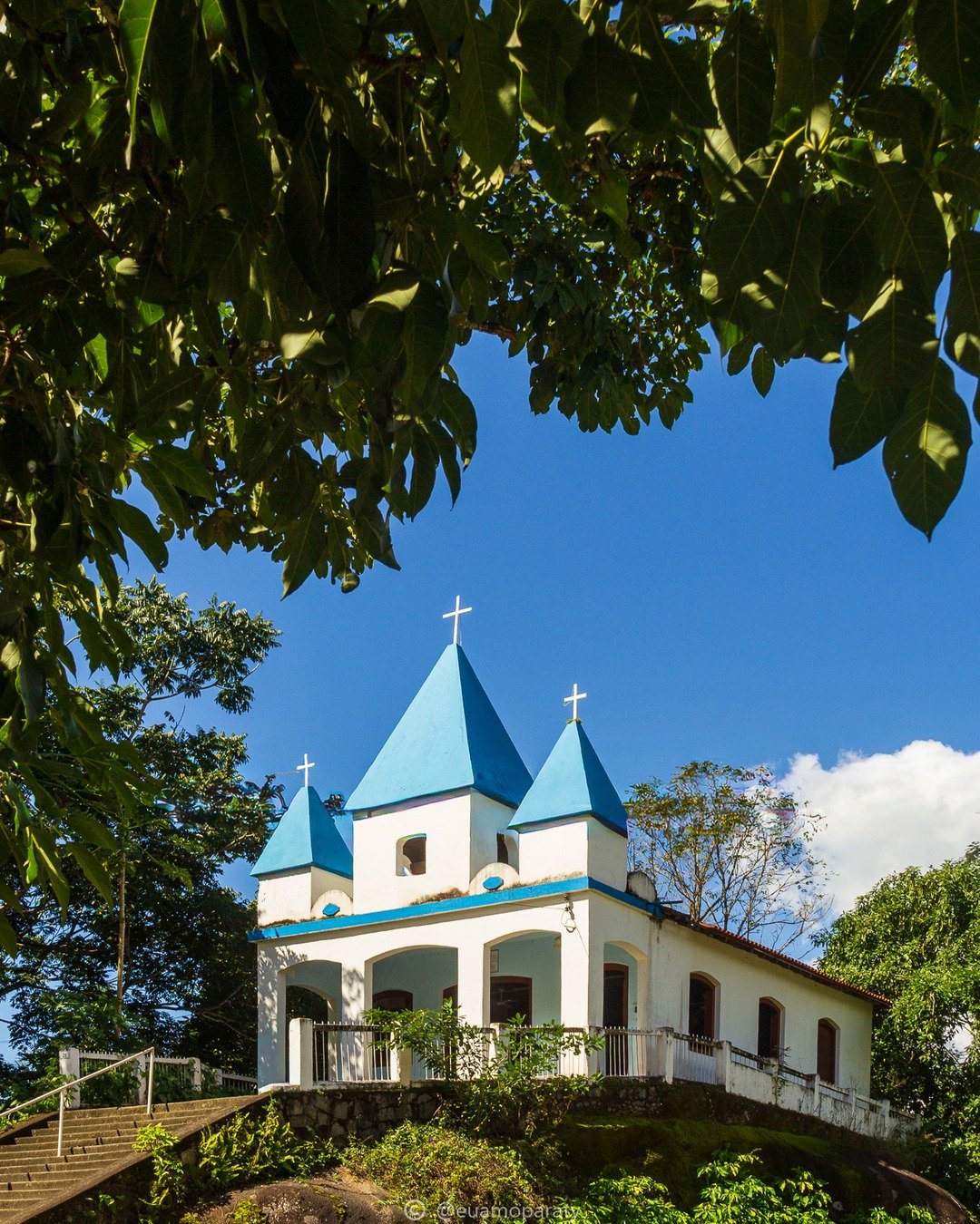 Igreja branca com telhados azuis e cruzes, vista entre folhagens verdes sob céu azul.