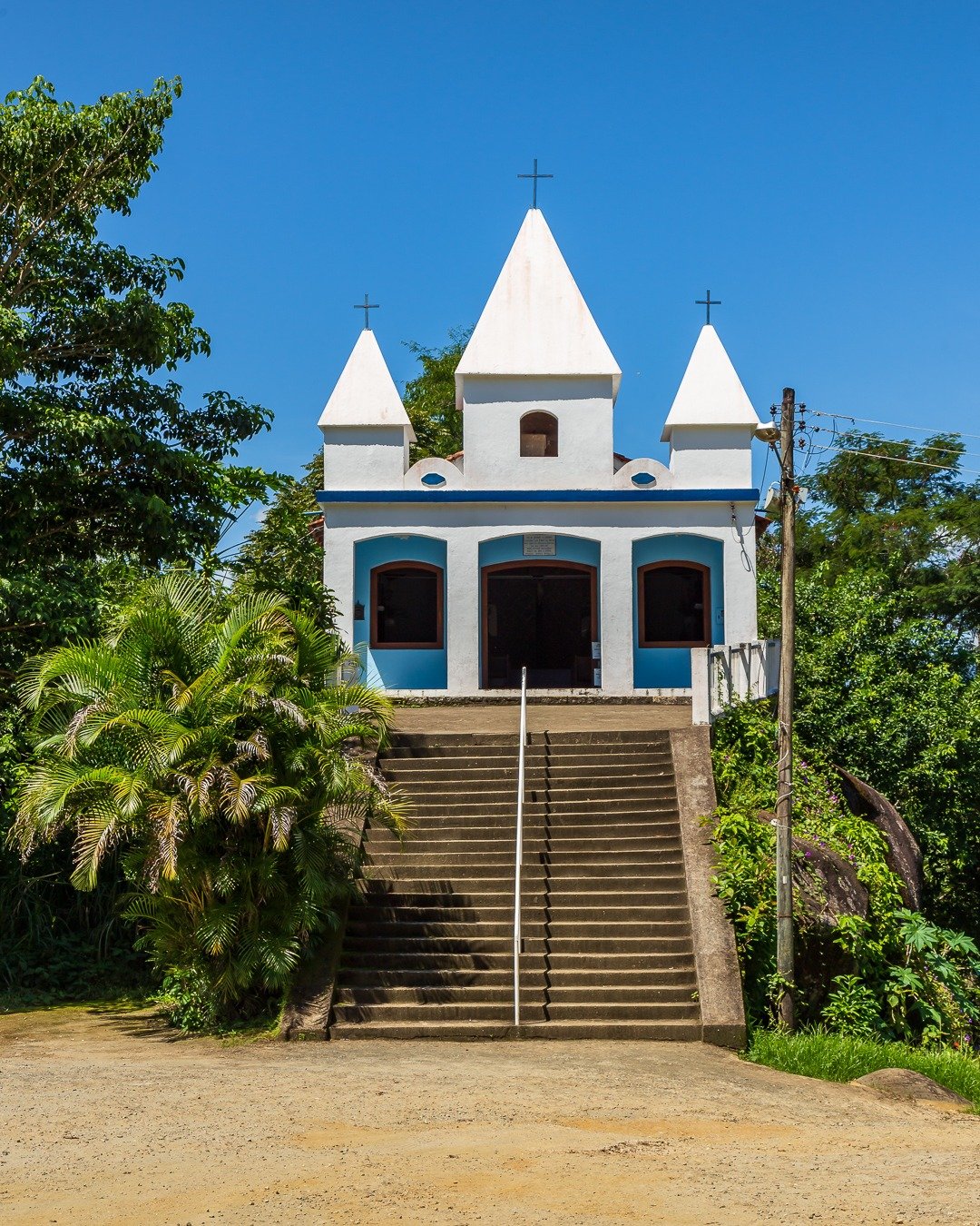 Capela branca com detalhes azuis no alto de uma escadaria, cercada por vegetação sob céu azul.