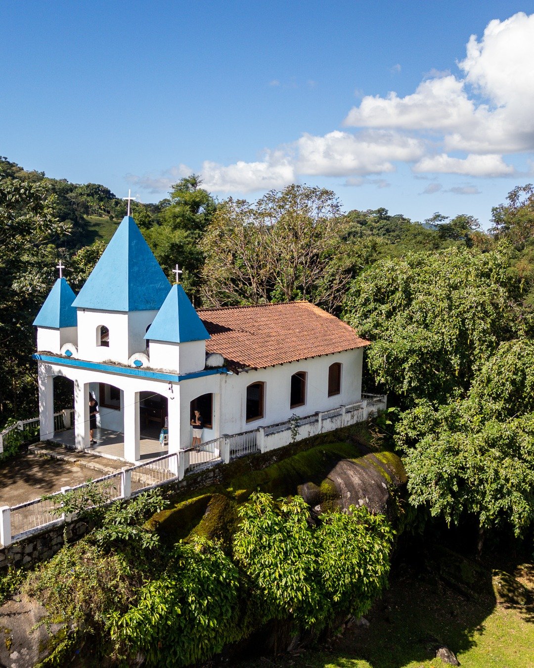 Vista aérea de uma capela branca com telhados azuis sobre um morro rochoso cercado por floresta, com escadaria de pedra.