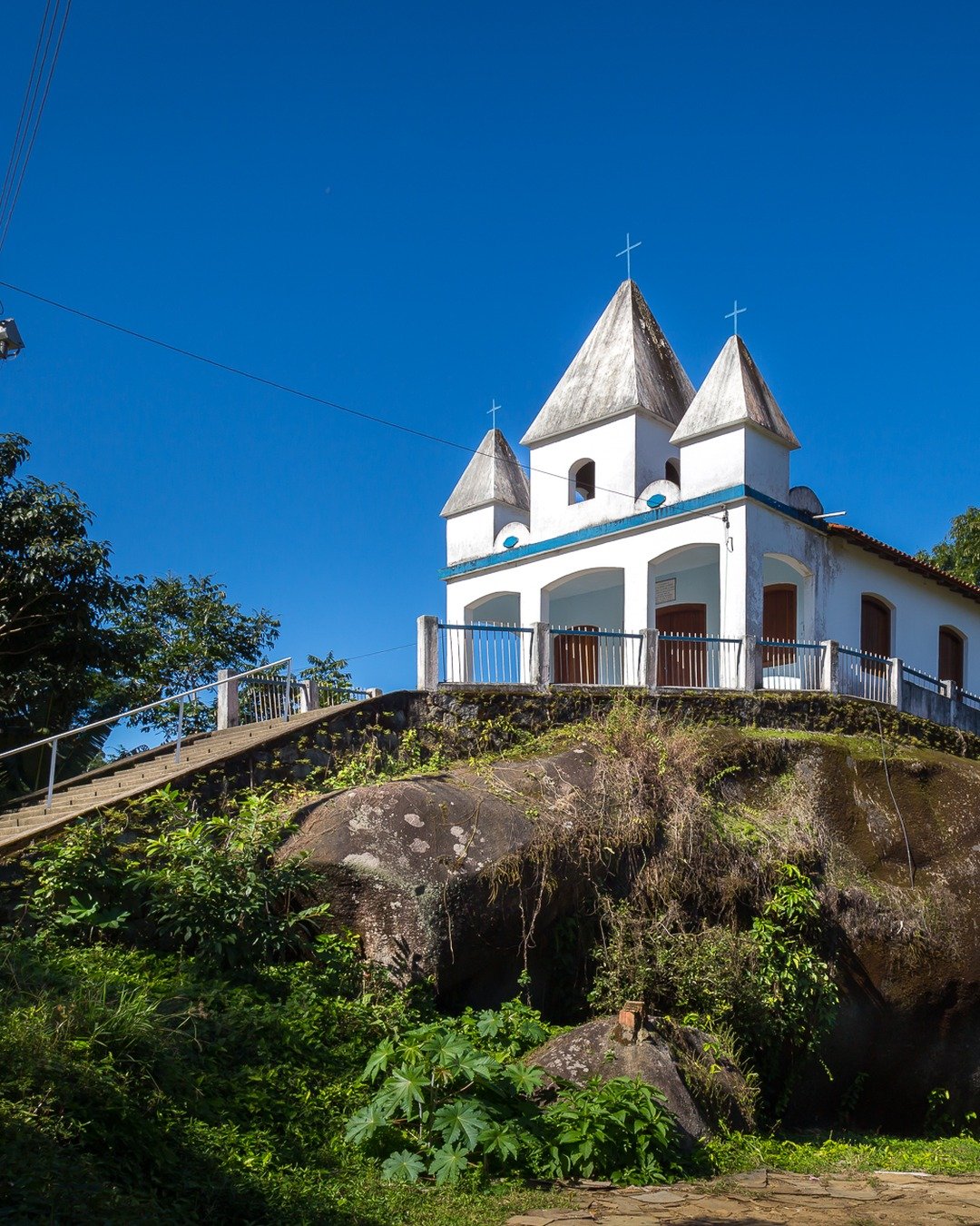 Igreja branca com três torres sobre uma grande rocha, cercada por vegetação e céu azul intenso.