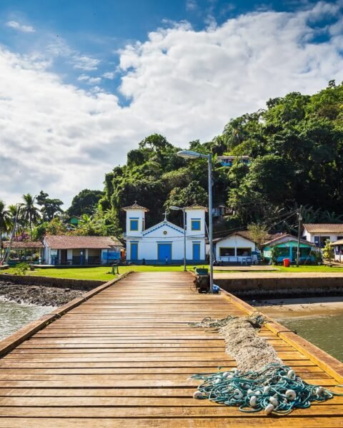 Pier de madeira conduz a uma pequena igreja branca de portas azuis, cercada por morros verdes e vegetação, sob céu azul com nuvens; redes de pesca aparecem no primeiro plano.