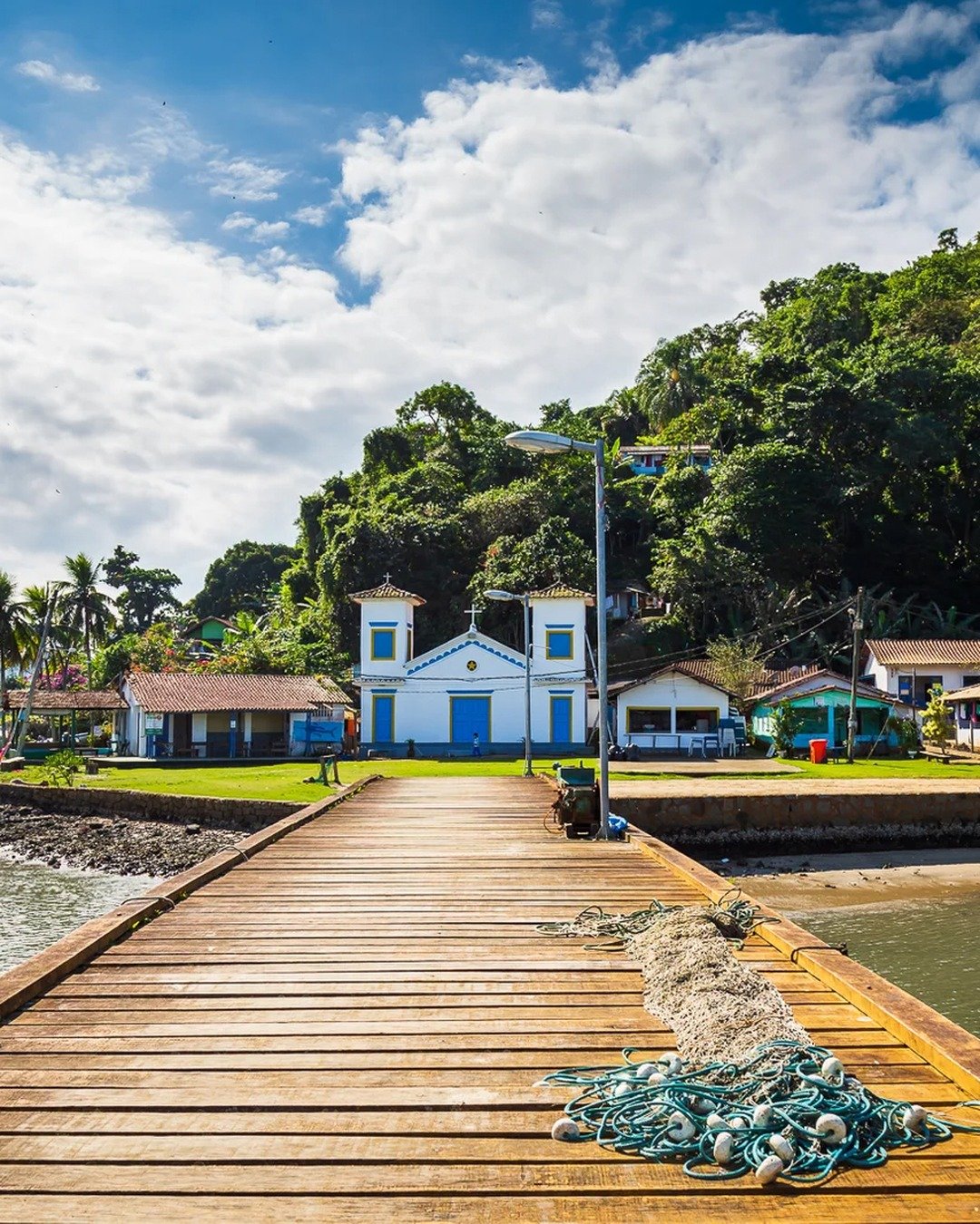 Pier de madeira conduz a uma pequena igreja branca de portas azuis, cercada por morros verdes e vegetação, sob céu azul com nuvens; redes de pesca aparecem no primeiro plano.