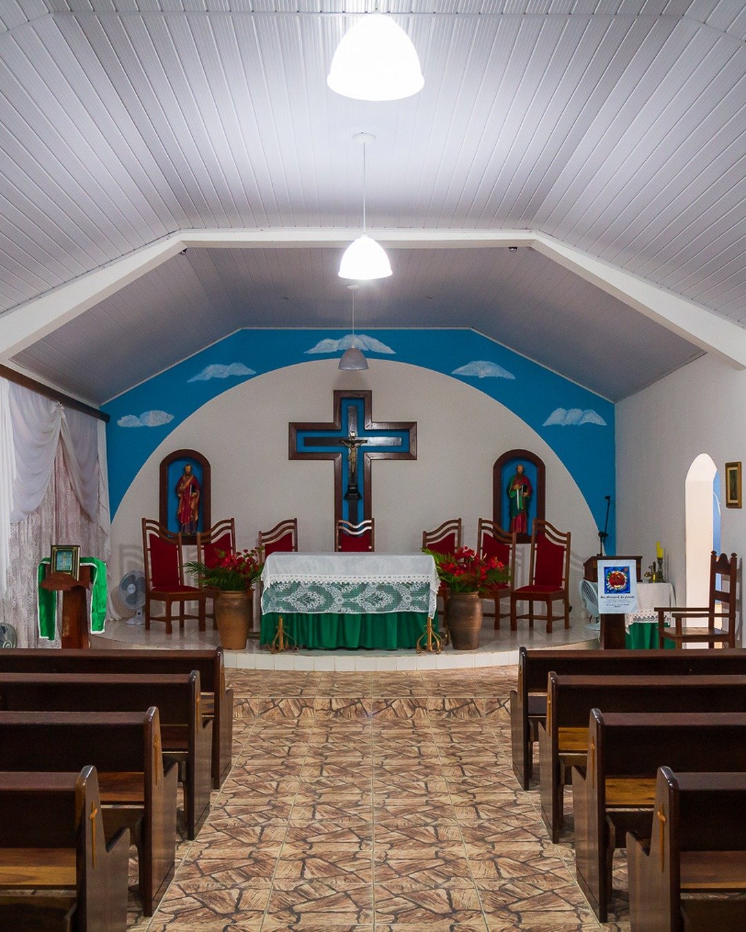 Interior de igreja com fileiras de bancos de madeira, altar central e cruz azul ao fundo.