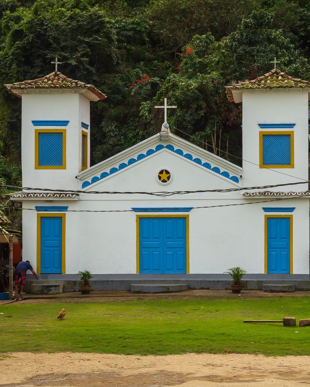 Igreja branca com detalhes azuis e amarelos, duas torres com cruzes, situada em meio a vegetação densa, com gramado e área de terra na frente. Ao lado direito, uma construção menor com mesas e cadeiras.