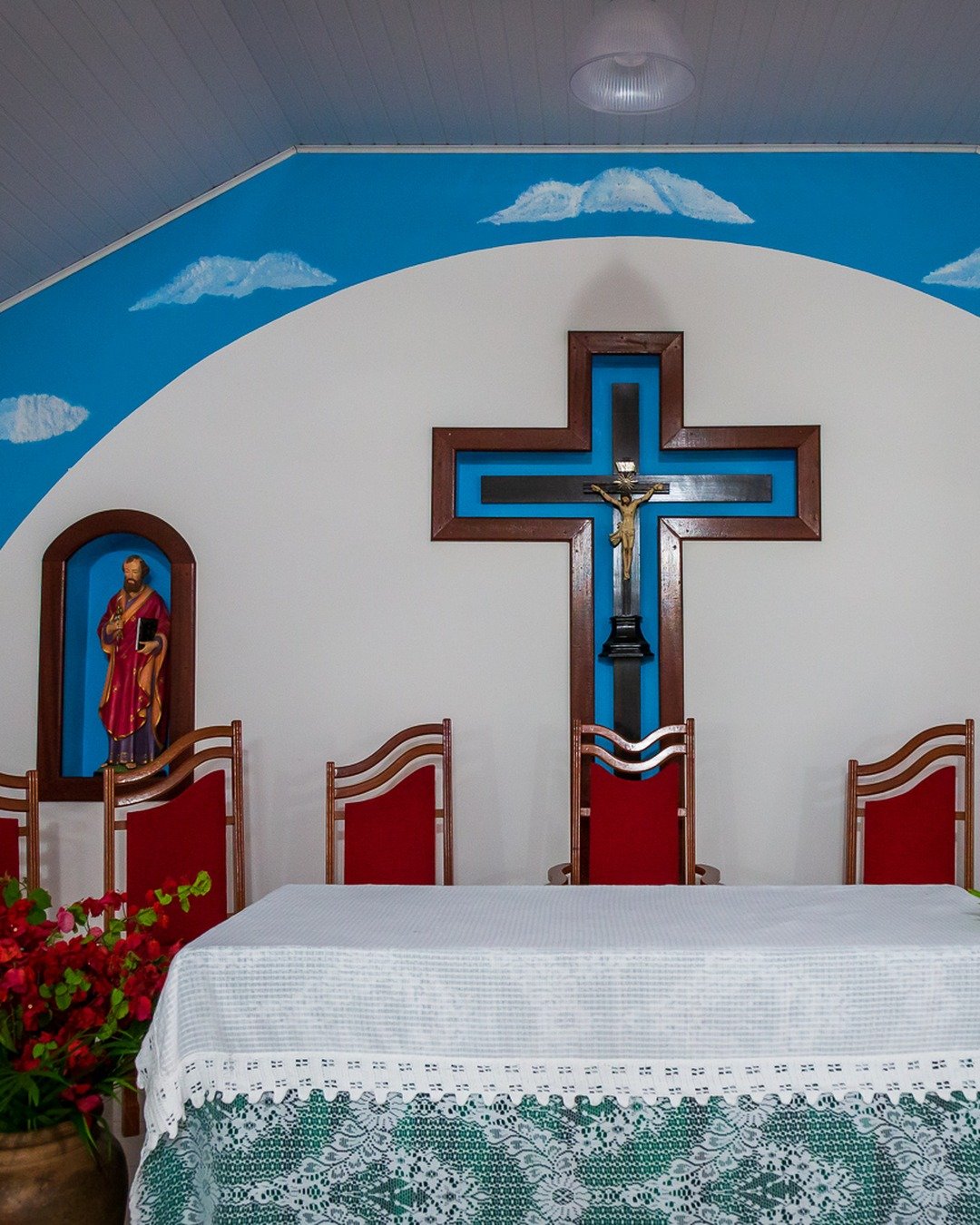 Interior de capela com altar coberto por toalha de renda, cruz central, nichos com santos e cadeiras vermelhas.