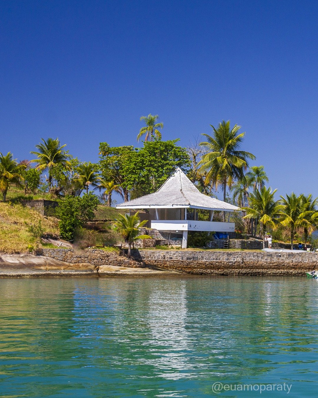 Vista de um ponto costeiro com um quiosque/pavilhão branco de telhado cônico sobre pier de pedra, cercado por palmeiras, com mar calmo esverdeado em primeiro plano e montanhas ao fundo sob céu azul limpo.