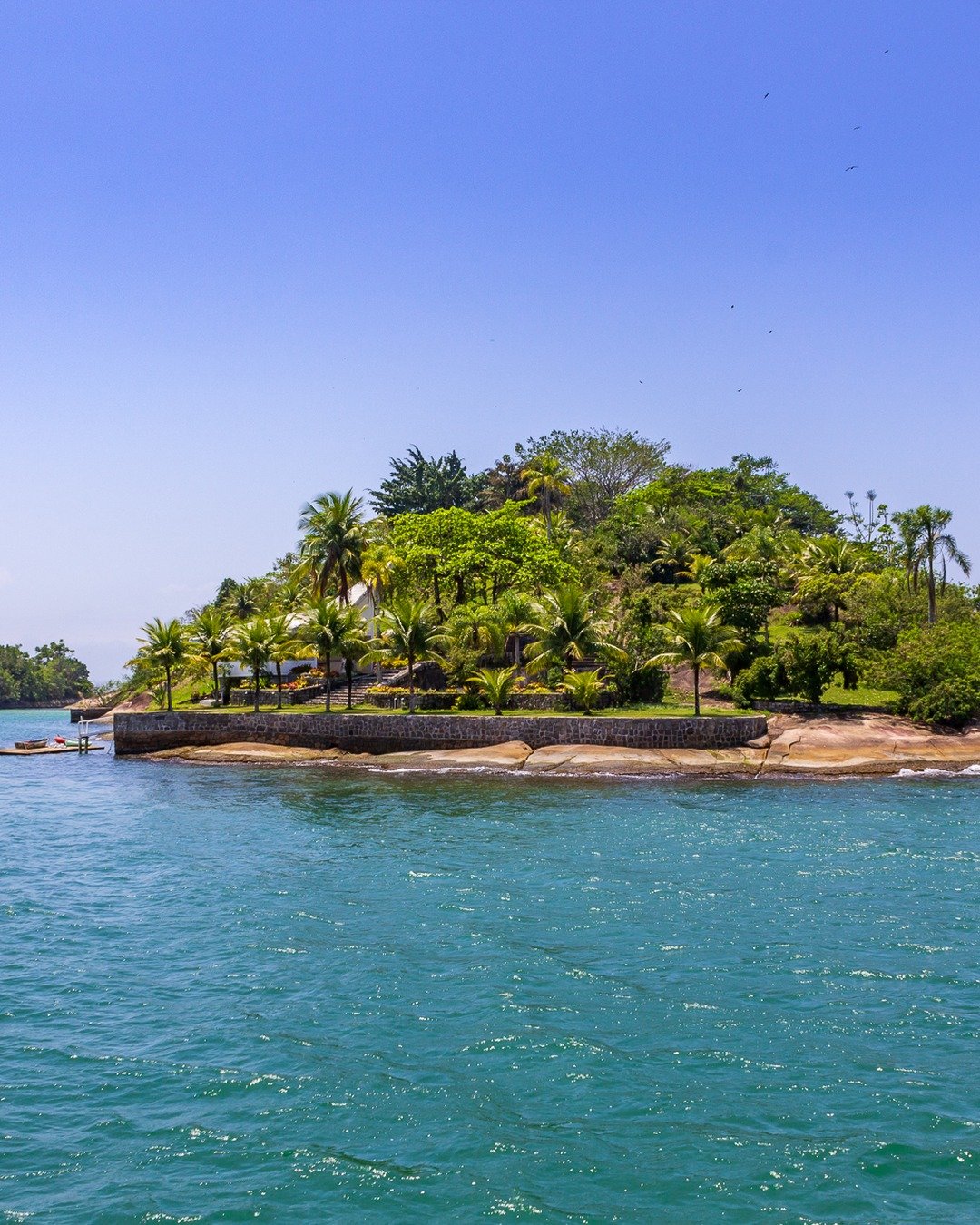 Ilha tropical com vegetação densa e palmeiras cercada por mar verde-turquesa, sob céu azul com poucas nuvens; outra porção de ilha aparece ao fundo à esquerda.