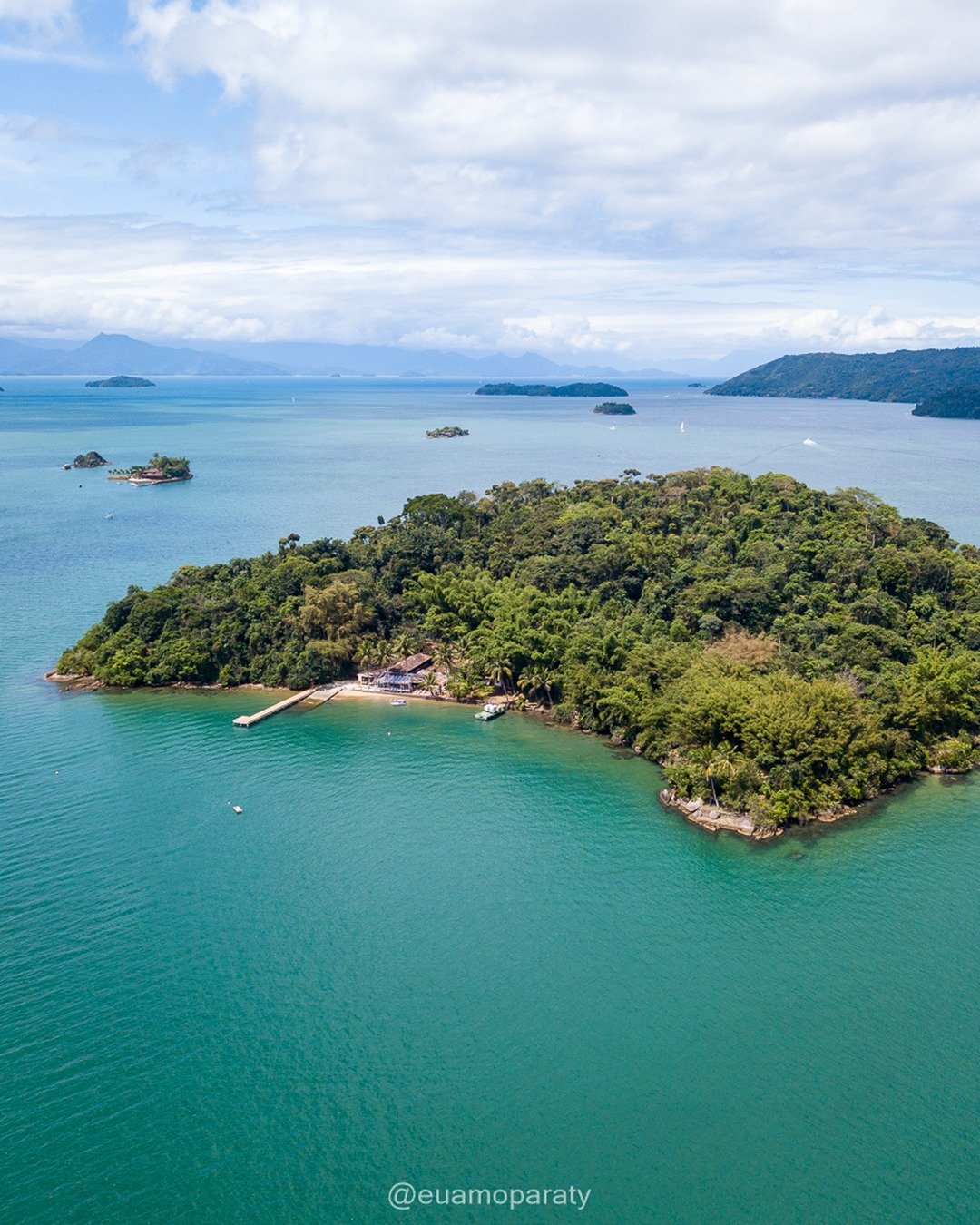 Vista aérea de uma ilha coberta por vegetação densa cercada por água verde-turquesa, com pequenas embarcações, ilhas ao fundo e montanhas no horizonte sob céu com nuvens.