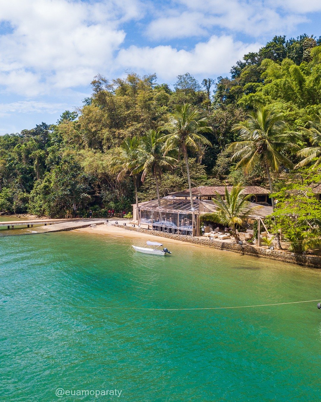 Vista elevada de uma enseada com água turquesa, praia pequena com restaurante à beira-mar, píer de madeira, embarcações e encosta de mata atlântica com palmeiras ao fundo.