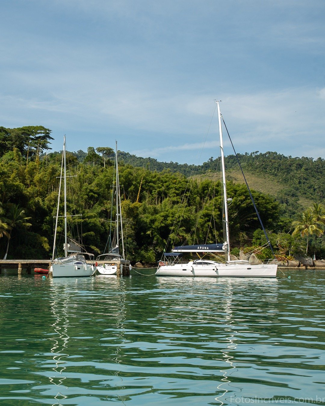 Baía de águas verdes e calmas com veleiros ancorados junto a um píer de madeira, tendo ao fundo encosta coberta por mata tropical densa e construções parcialmente ocultas pela vegetação.