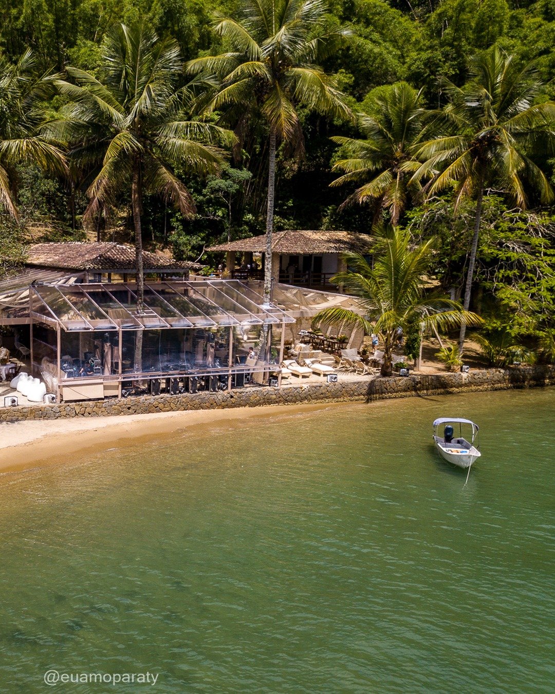 Vista aérea de um restaurante rústico à beira-mar, cercado por palmeiras e mata fechada, com pequena faixa de areia, pedras e uma lancha ancorada na água esverdeada.