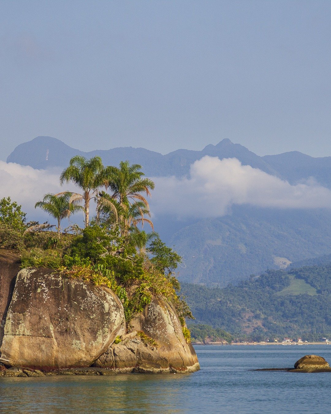 Ponta rochosa coberta de vegetação e palmeiras à beira-mar, com montanhas ao fundo sob nuvens e um pequeno barco na água.