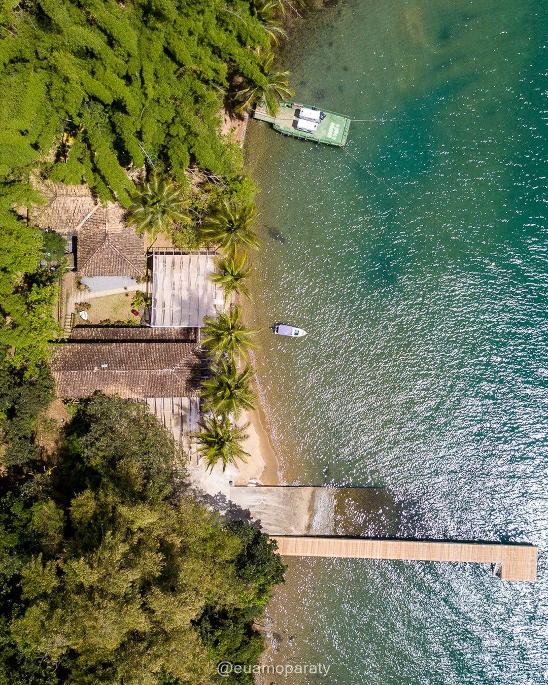 Vista aérea de uma propriedade à beira-mar cercada por vegetação densa, com praia estreita, píer de madeira adentrando a água e duas embarcações ancoradas.