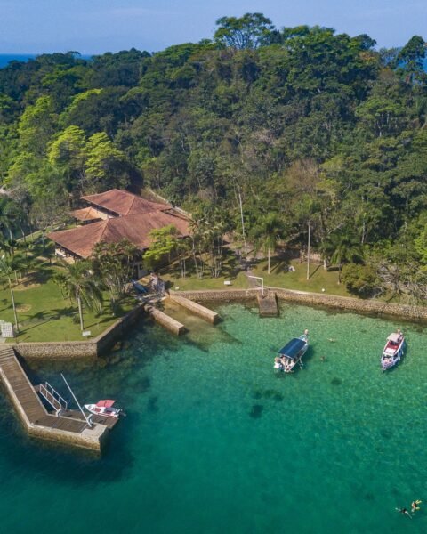 Vista aérea de uma enseada com águas turquesa, casa de telhado vermelho sobre ponta gramada, cais de pedra com barcos ancorados e morro coberto por mata atlântica.