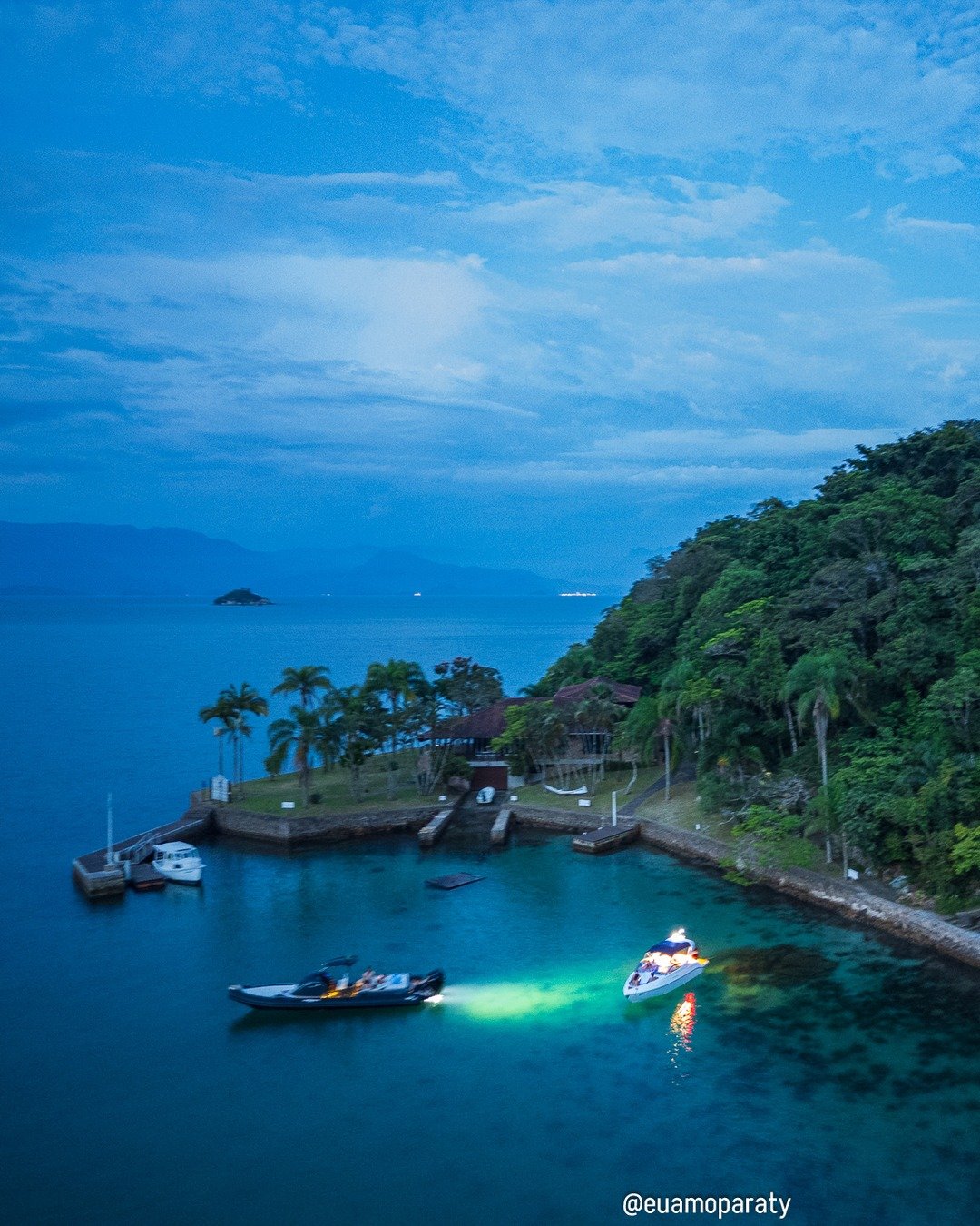 Vista aérea ao anoitecer de uma enseada com dois barcos iluminados na água, um deles com luz verde, próximos a uma pequena ilha com vegetação densa e construções, sob céu azul com nuvens.