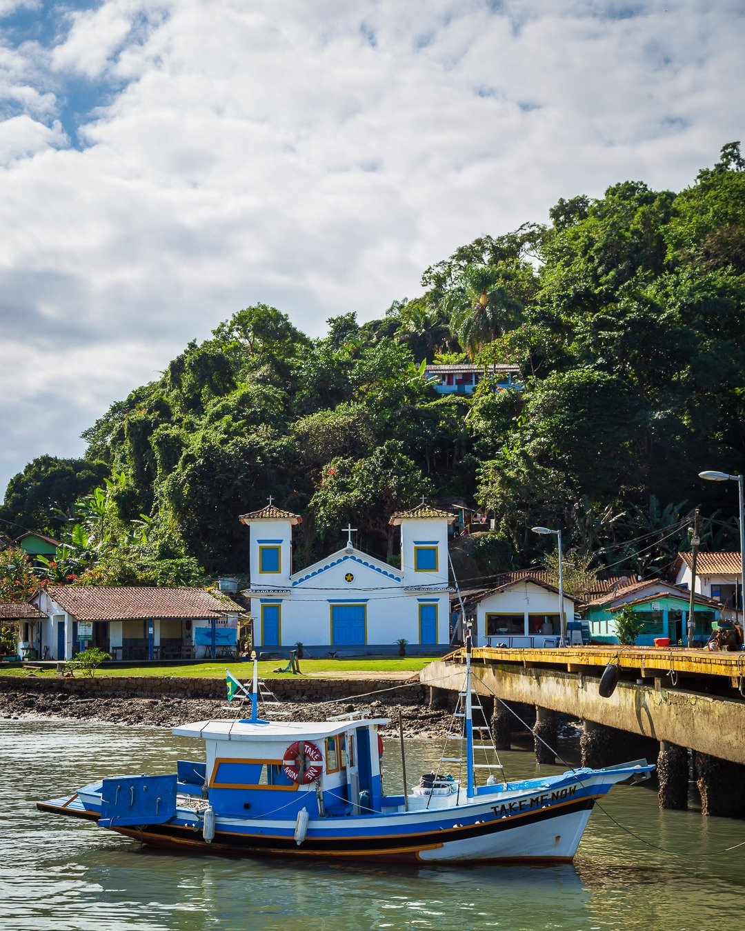 Barco de pesca azul e branco ancorado junto a um cais de concreto, com uma pequena igreja branca de detalhes azuis na margem e uma encosta densamente arborizada ao fundo sob céu parcialmente nublado.