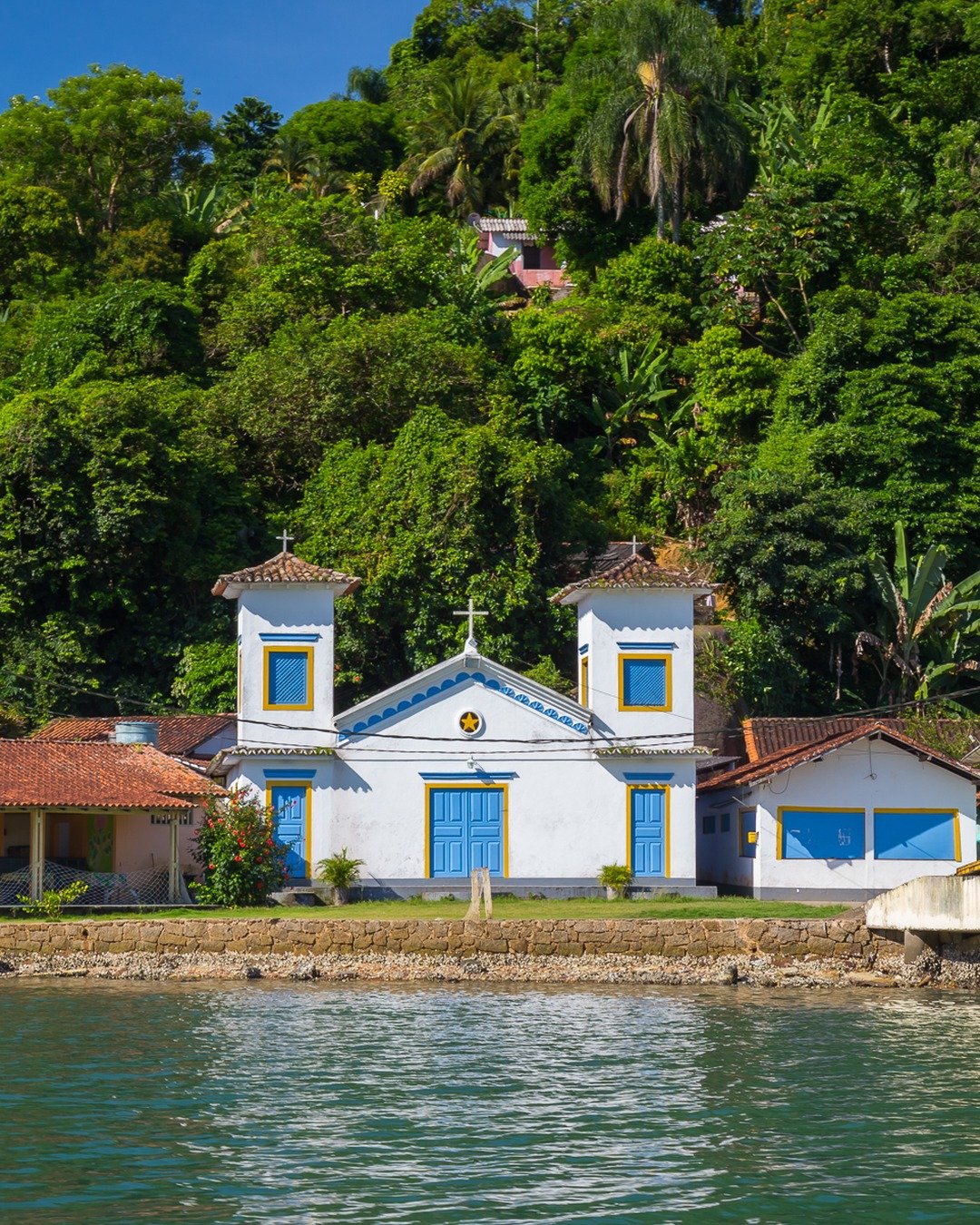 Igreja branca com detalhes azuis à beira da água, tendo ao fundo um morro densamente coberto por vegetação tropical e o céu azul no topo.